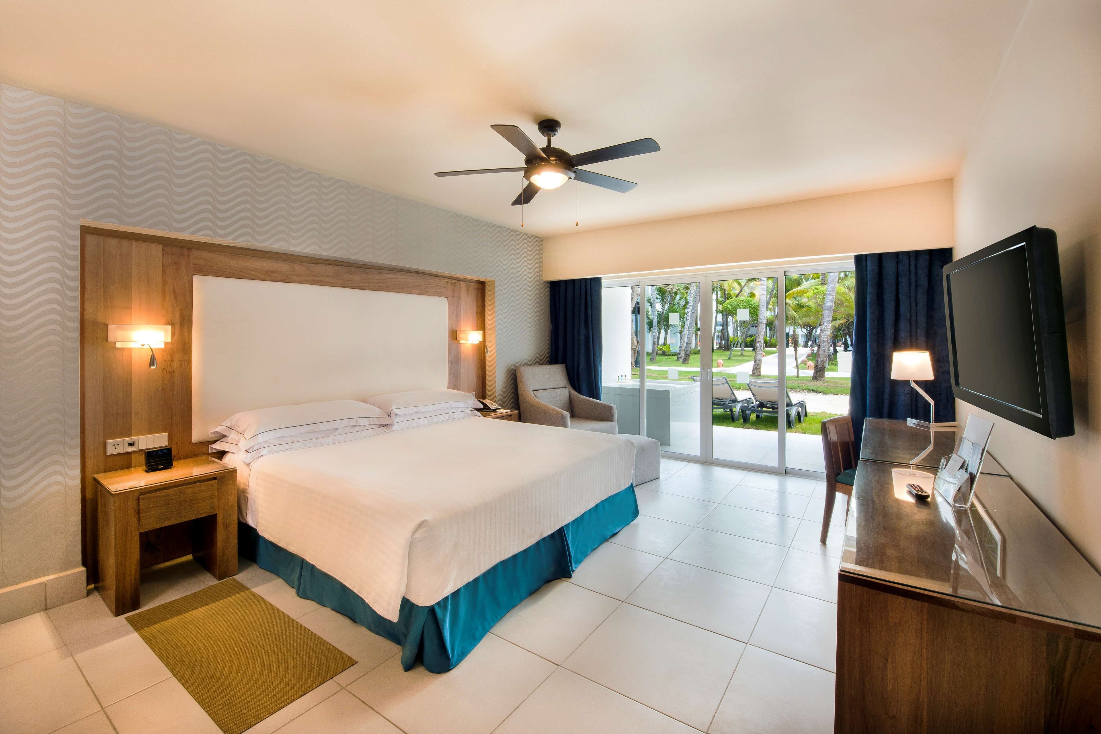 A hotel room with a large bed, a desk, a television, a ceiling fan, and a view of the outdoors.