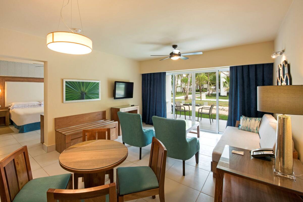 A hotel room with a bed, a desk, a television, a balcony with chairs, and a ceiling fan.