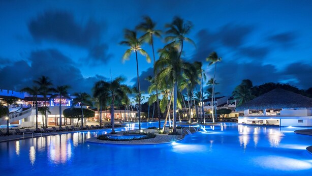 Ein Poolbereich eines Resorts mit Palmen, einem Gebäude mit Reetdach und einem gut beleuchteten Schwimmbad bei Einbruch der Dunkelheit.