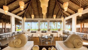 Ein Restaurant mit einem Reetdach, weißem Mobiliar und Blick auf das Meer.