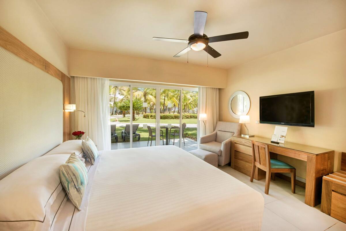 A hotel room with a large bed, a desk with a chair, a television, and a view of palm trees through the window.