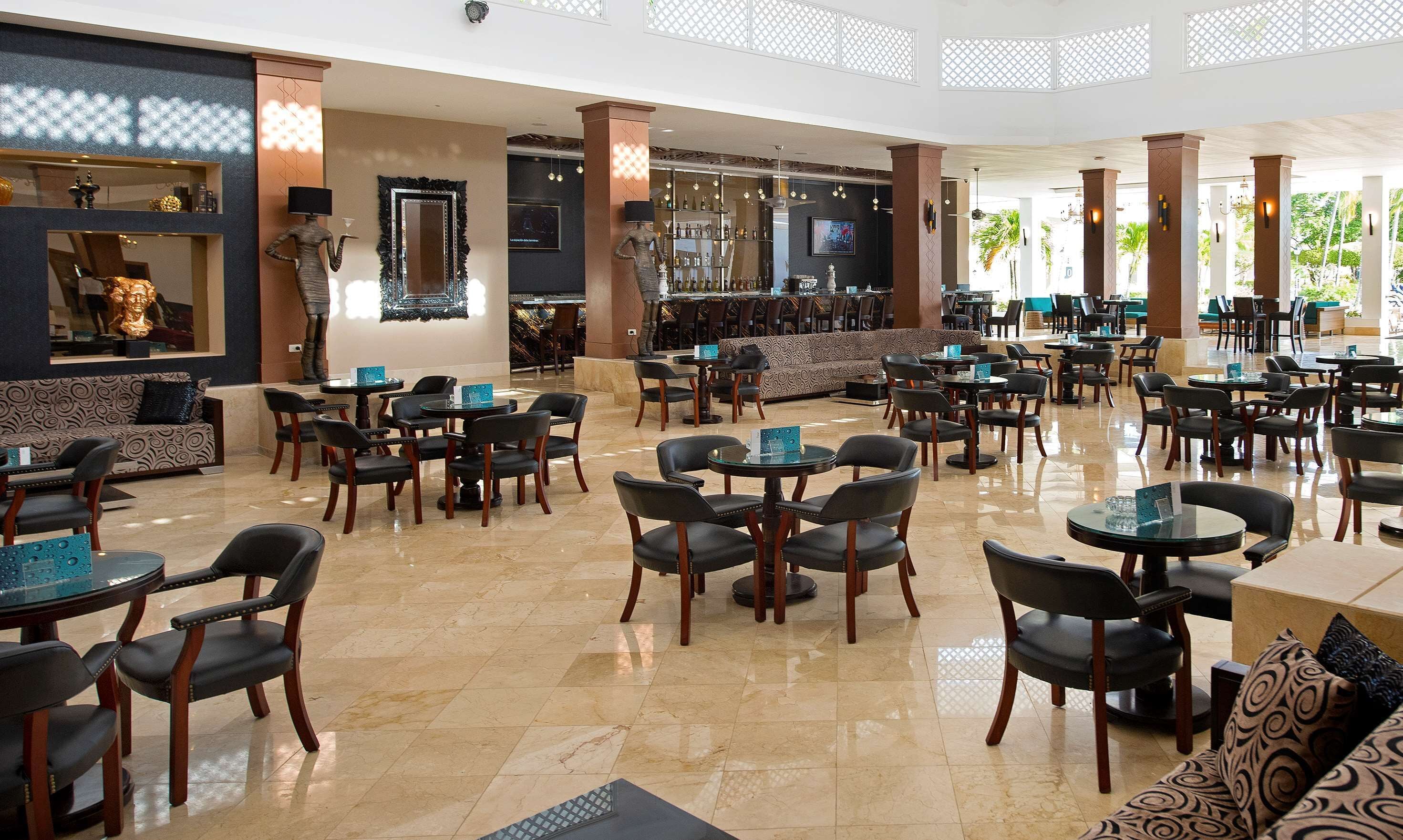 Un amplio vestíbulo de hotel con varias zonas de estar, incluidas mesas redondas y sofás, y una zona de bar con un espejo grande y elementos decorativos.