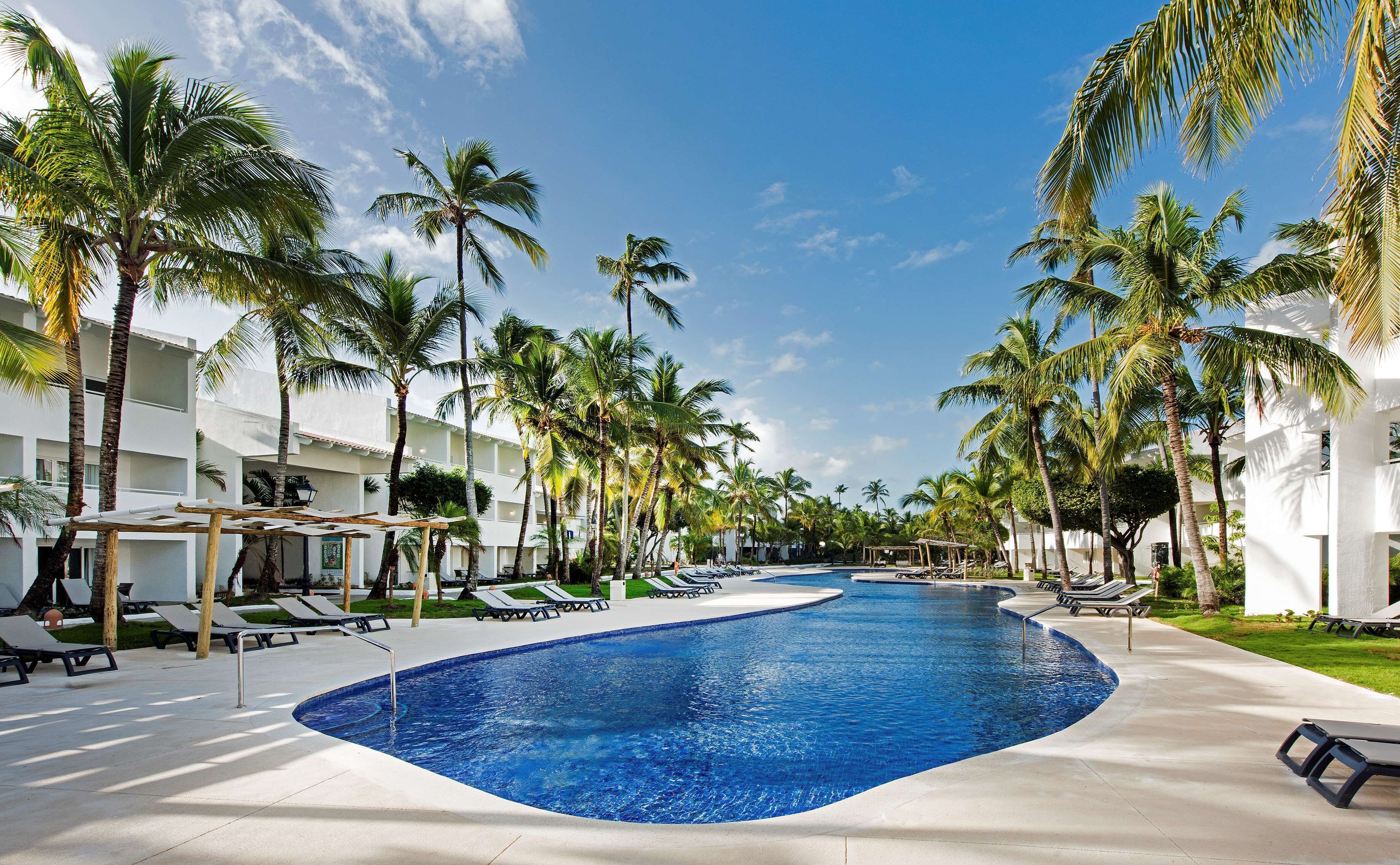 A swimming pool surrounded by palm trees and lounge chairs, with a clear blue sky above.