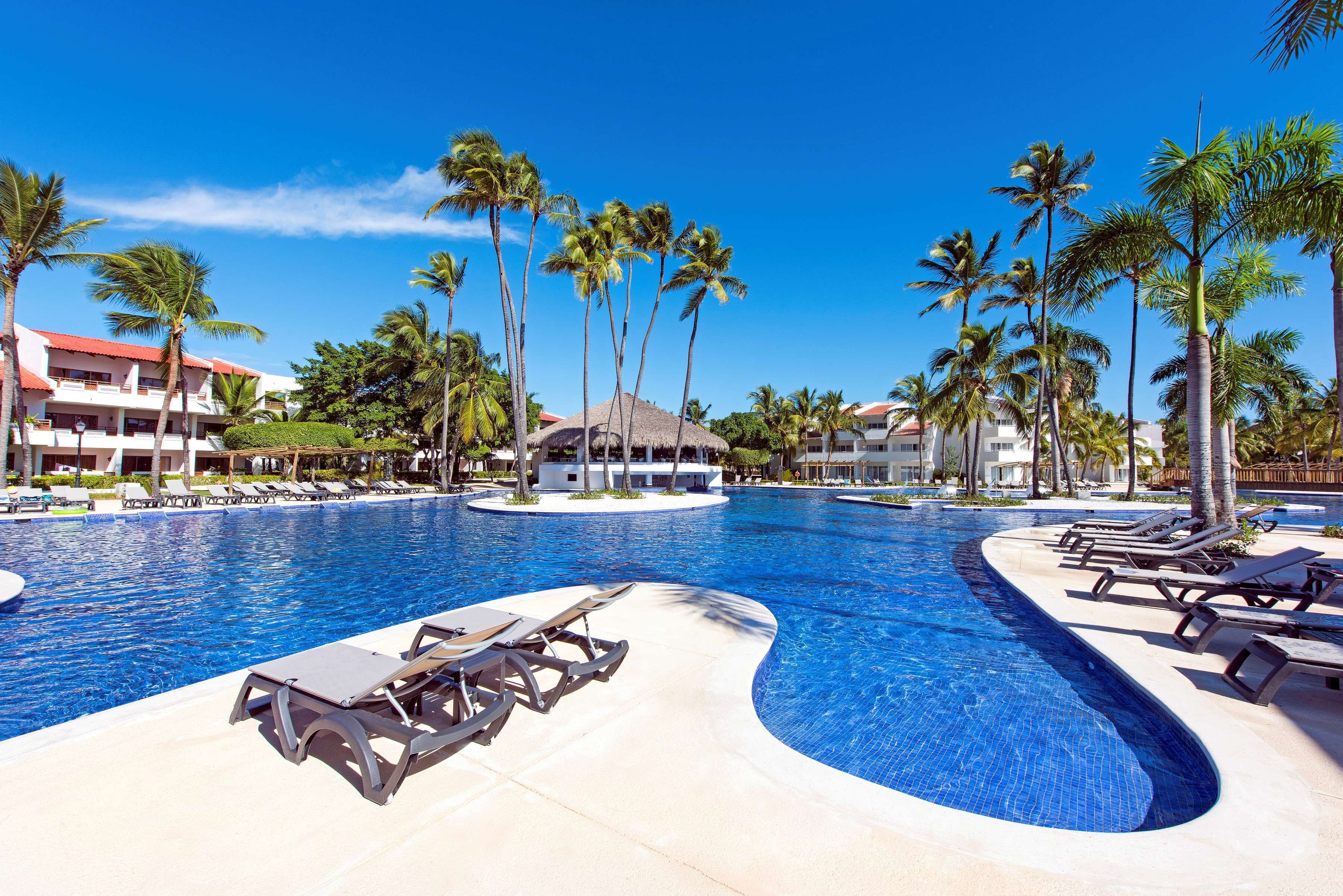 Een zwembad van een resort met ligbedden, palmbomen en gebouwen van het resort op de achtergrond.