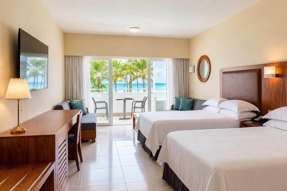 A hotel room with two beds, a desk, a chair, a TV, and a balcony with a view of palm trees and the ocean.