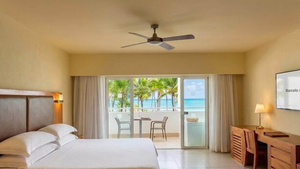 A hotel room with a bed, a desk, a chair, a ceiling fan, and a view of the ocean.
