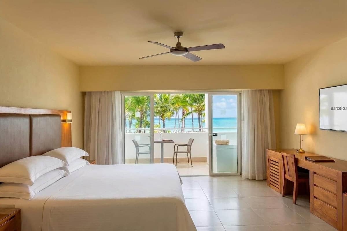 A hotel room with a bed, a desk, a chair, a ceiling fan, and a view of the ocean.
