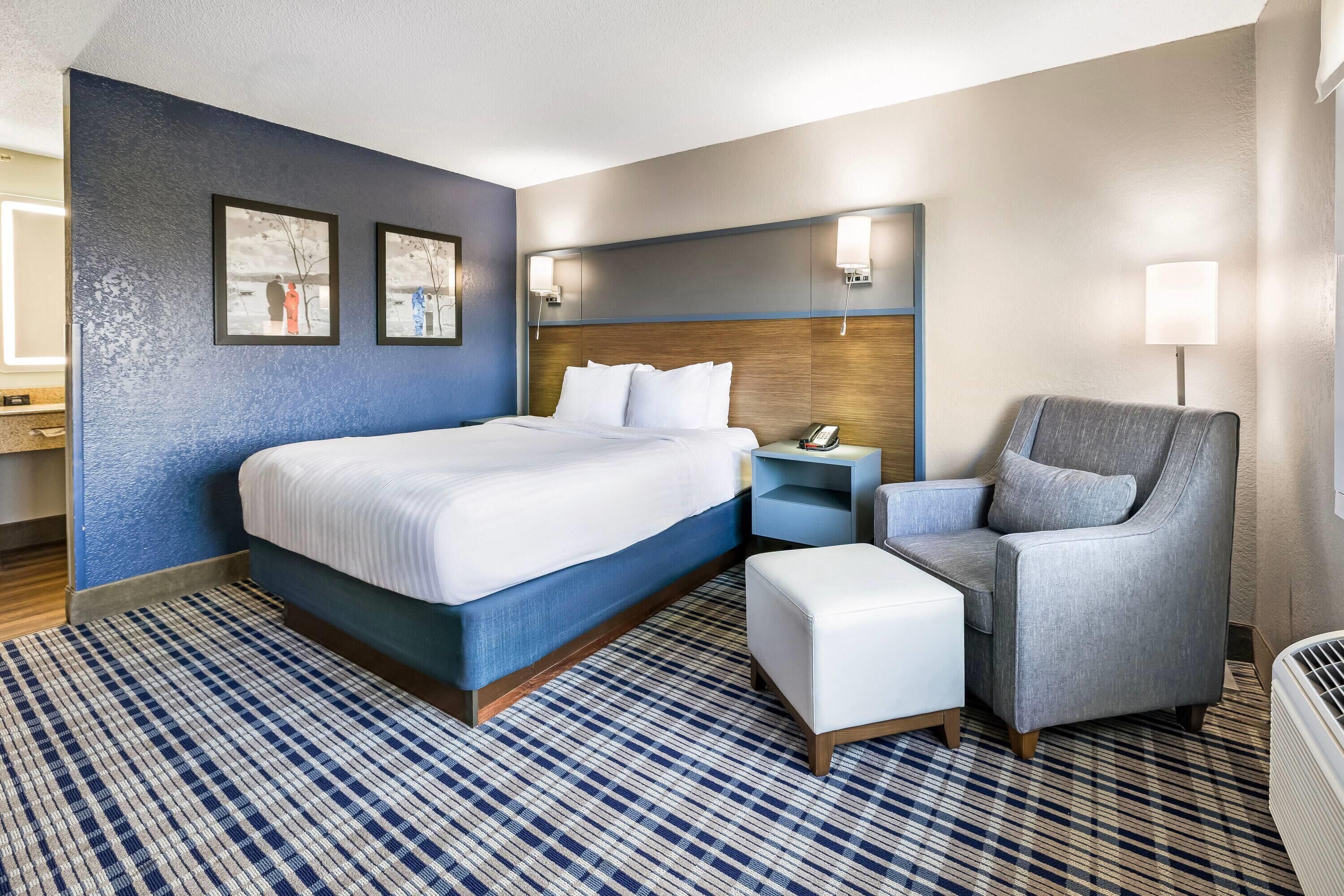 A hotel room with a large bed, a bedside table with a lamp, a chair, and a patterned rug.