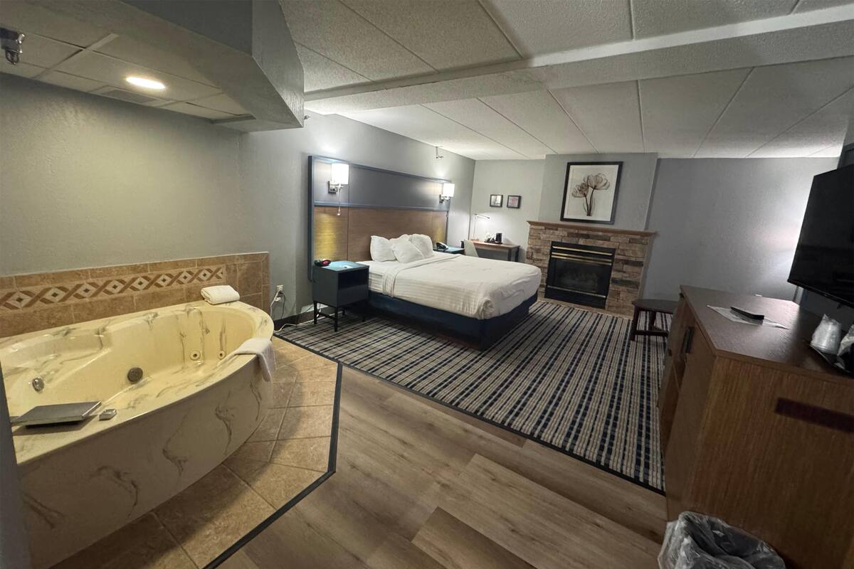 A hotel room with a large bed, a separate bathroom with a jacuzzi, a desk, and a fireplace.