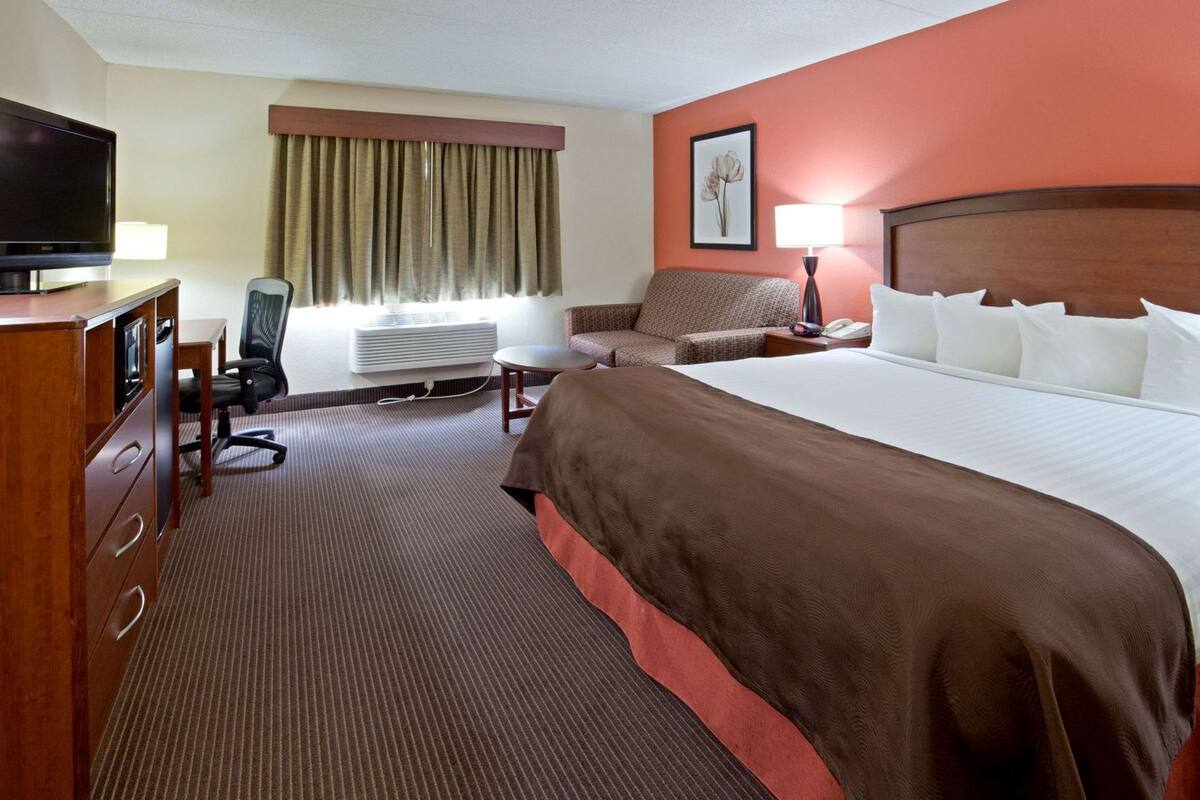 A hotel room with a bed, a desk with a television, a chair, a lamp, and a window with curtains.