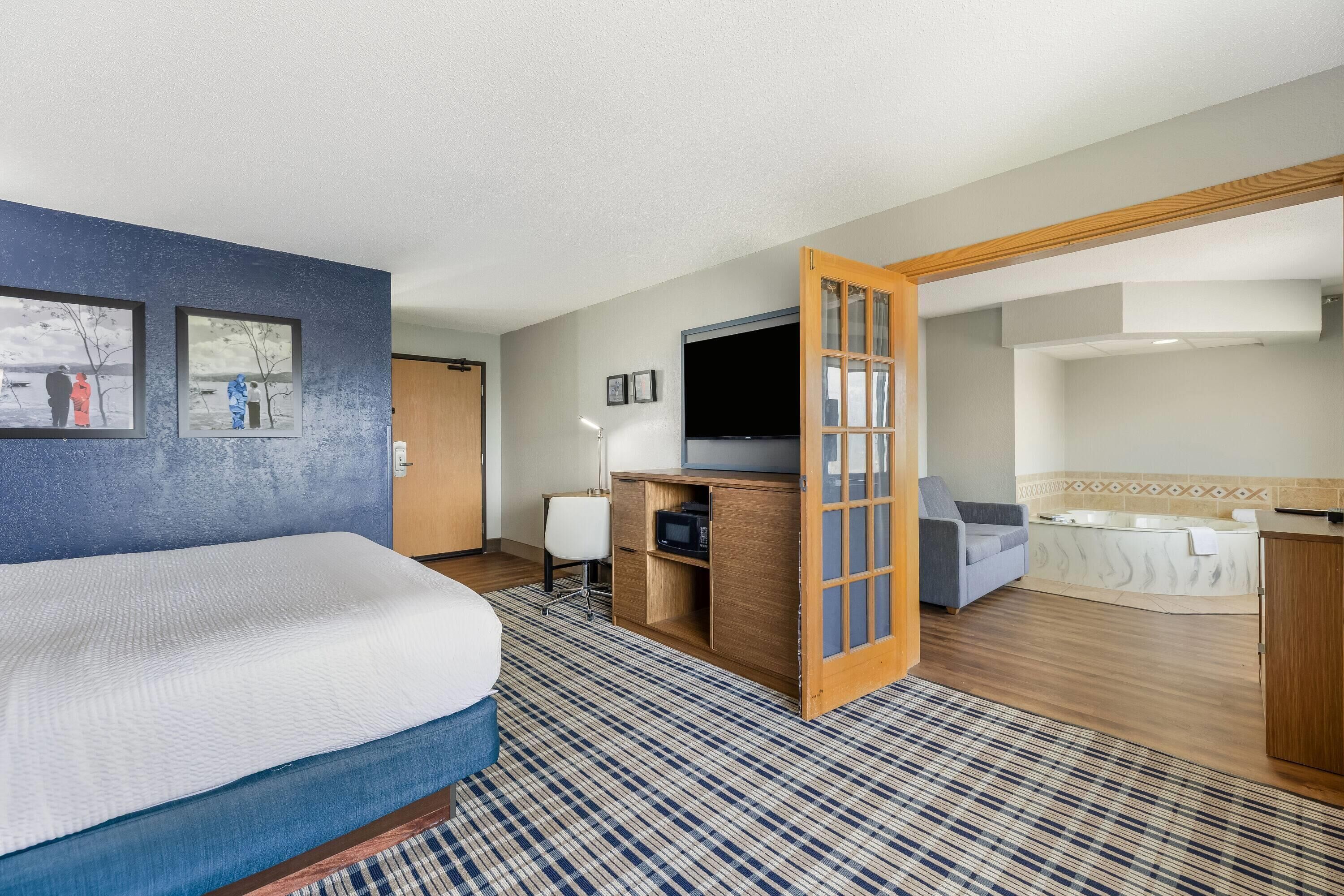 Habitación de hotel con cama, escritorio, televisión y baño con bañera, además de una puerta que da a otra habitación.