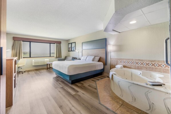 A hotel room with a bed, a desk, a chair, a window with curtains, a radiator, a wall-mounted picture, a floor lamp, and a bathtub. - AmericInn by Wyndham Albert Lea (Albert Lea)