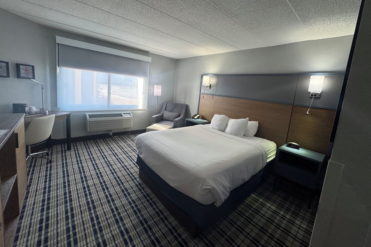 A hotel room with a large bed, a desk with a chair, a window, and a bedside table with a lamp.