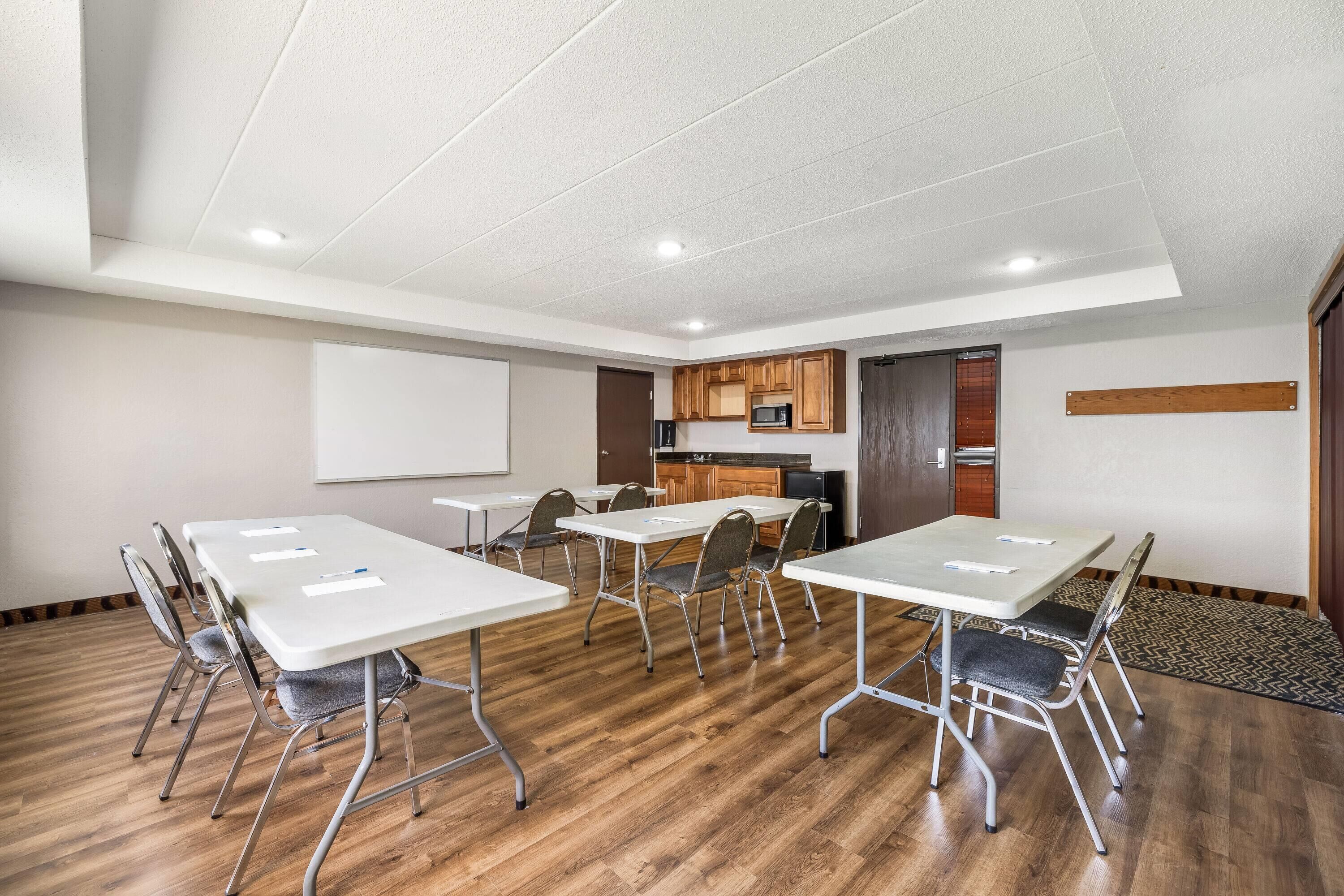 A conference room with white tables and chairs, a whiteboard, and a kitchen area with wooden cabinets.