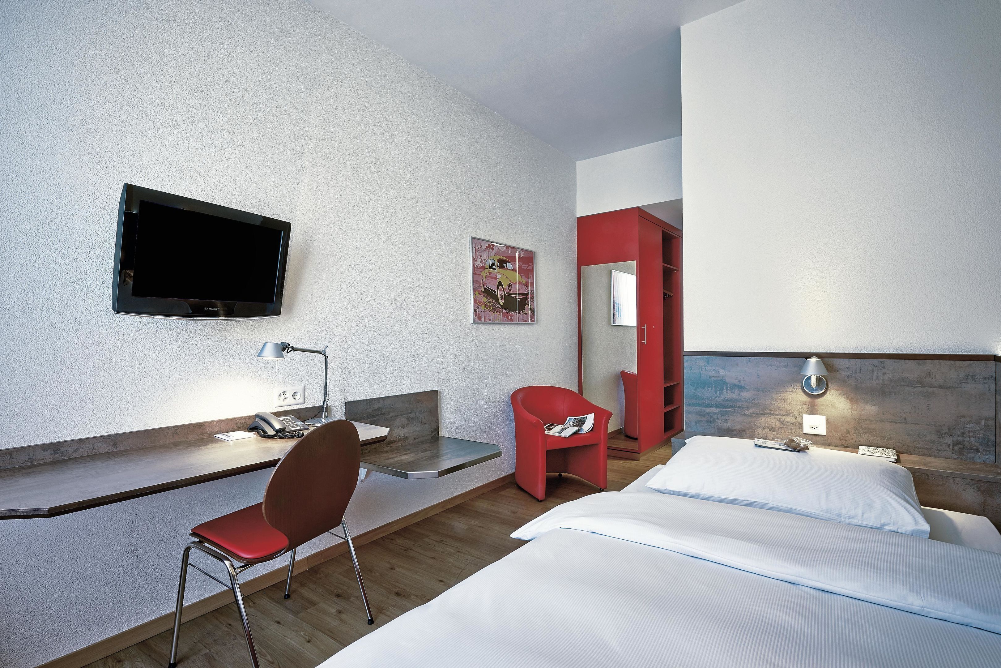 Habitación de hotel con cama, escritorio con silla, televisor, silla roja y lámpara de pared.