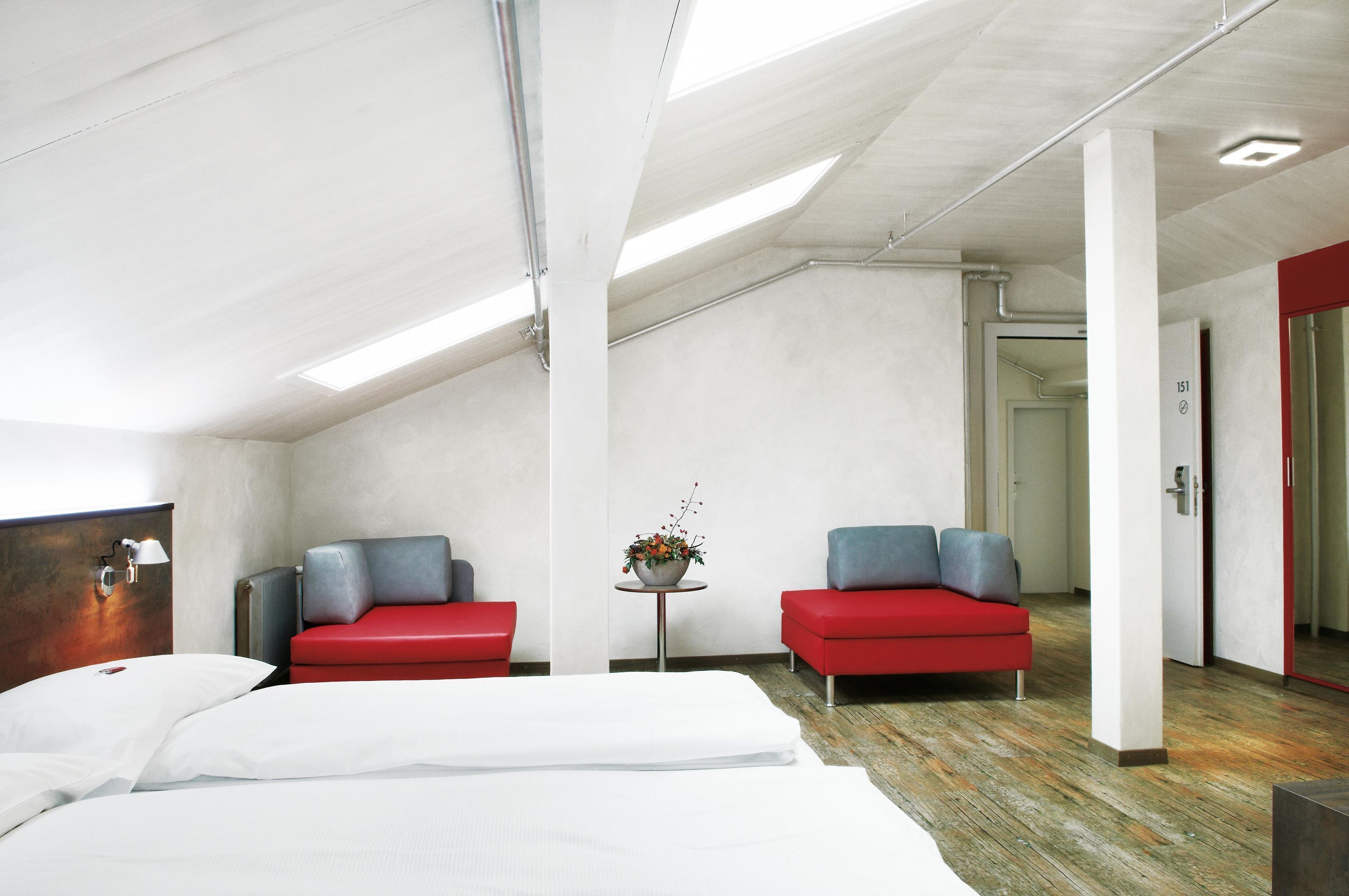 A modern hotel room with a large bed, two red sofas, a small table with a flower arrangement, and a visible door leading to another room.
