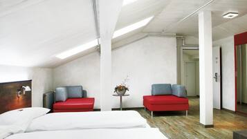 A modern hotel room with a large bed, two red sofas, a small table with a flower arrangement, and a visible door leading to another room.