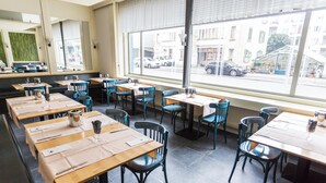 En restaurant med flere borde og stole, store vinduer med udsigt til gaden, og en grøn væg i baggrunden.