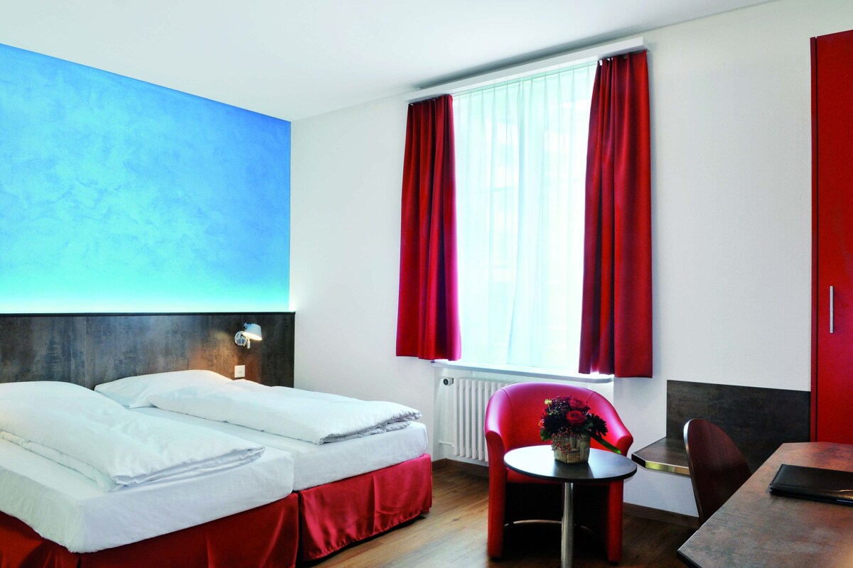 Una camera d'albergo con un letto, un tavolino con un vaso, una sedia, una finestra con tende e una porta rossa.