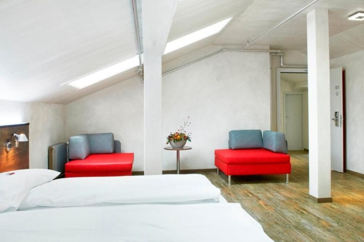 A hotel room with a high ceiling, skylights, a bed, two red sofas, a small table with a flower arrangement, and a door labeled '153'.