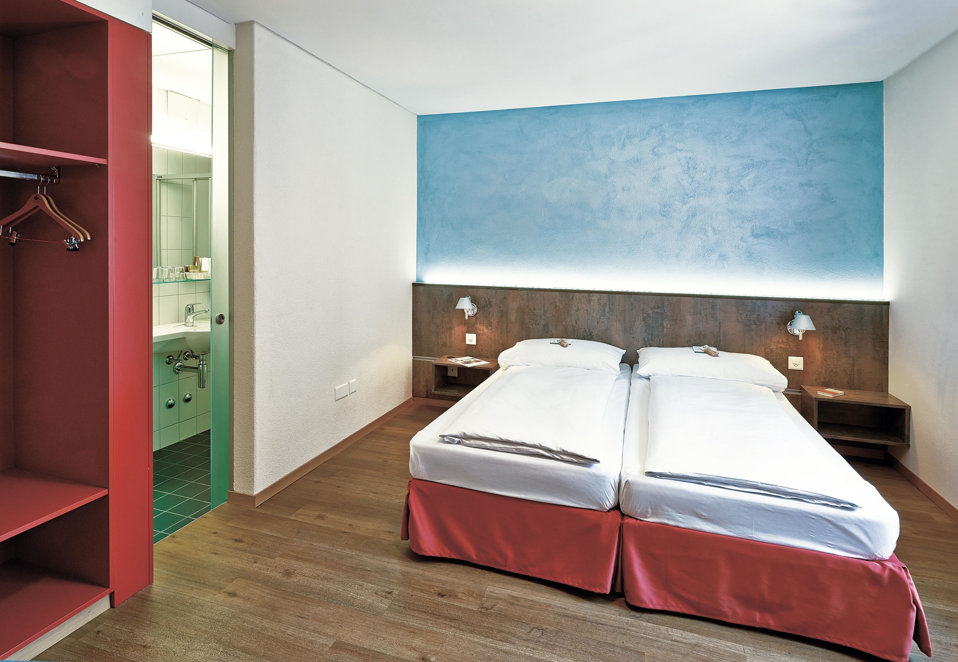 A hotel room with two beds, a wooden floor, a red wardrobe, and a bathroom visible through an open door.