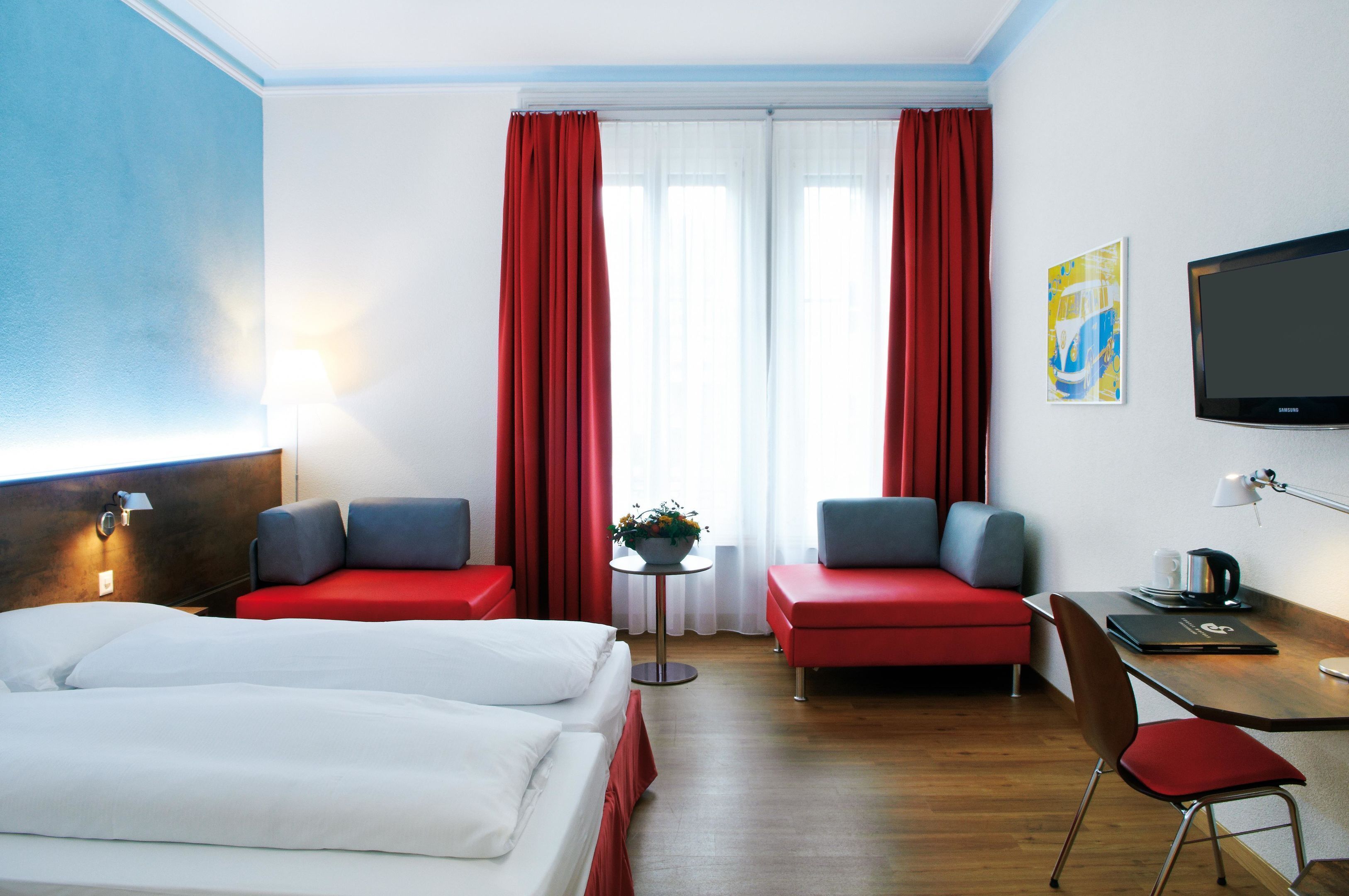 Habitación de hotel con una cama, dos sofás rojos, un escritorio con una silla, un televisor y una ventana con cortinas.