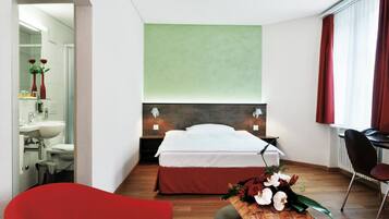 Une chambre d’hôtel avec un lit, un bureau, une chaise, un fauteuil rouge, une salle de bain avec un lavabo et un miroir, et un mur vert.