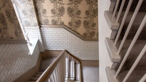 Un escalier avec des balustrades blanches et une main courante en bois, menant à un palier doté d’un mur à motifs et d’un sol carrelé de carreaux blancs.