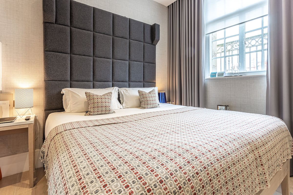 A hotel room with a large bed, a headboard, a bedside table with a lamp, and a window with curtains.
