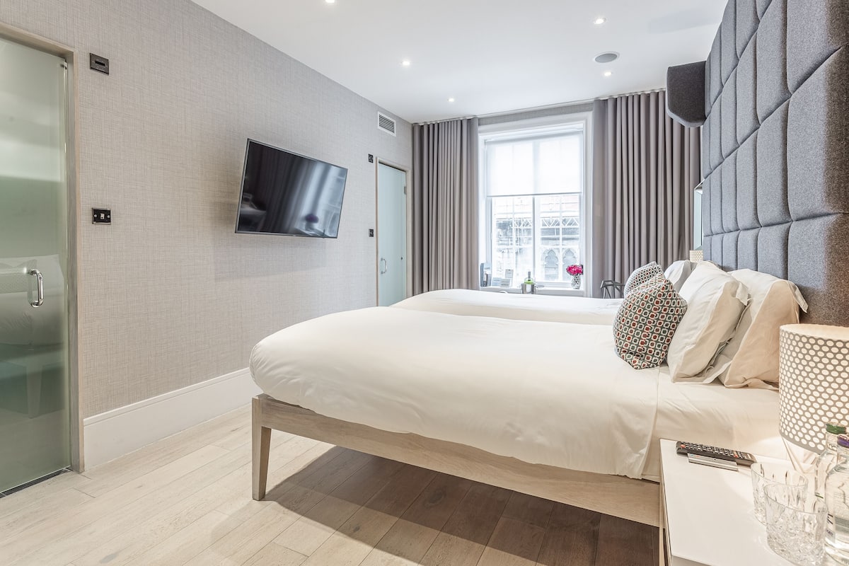A modern hotel room with a large bed, a flat-screen TV mounted on the wall, a glass shower enclosure, and a large window with curtains.