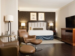 Quarto de hotel com cama, duas luminárias de cabeceira, televisão, armário de madeira, poltrona de couro marrom e uma mesinha com abajur.