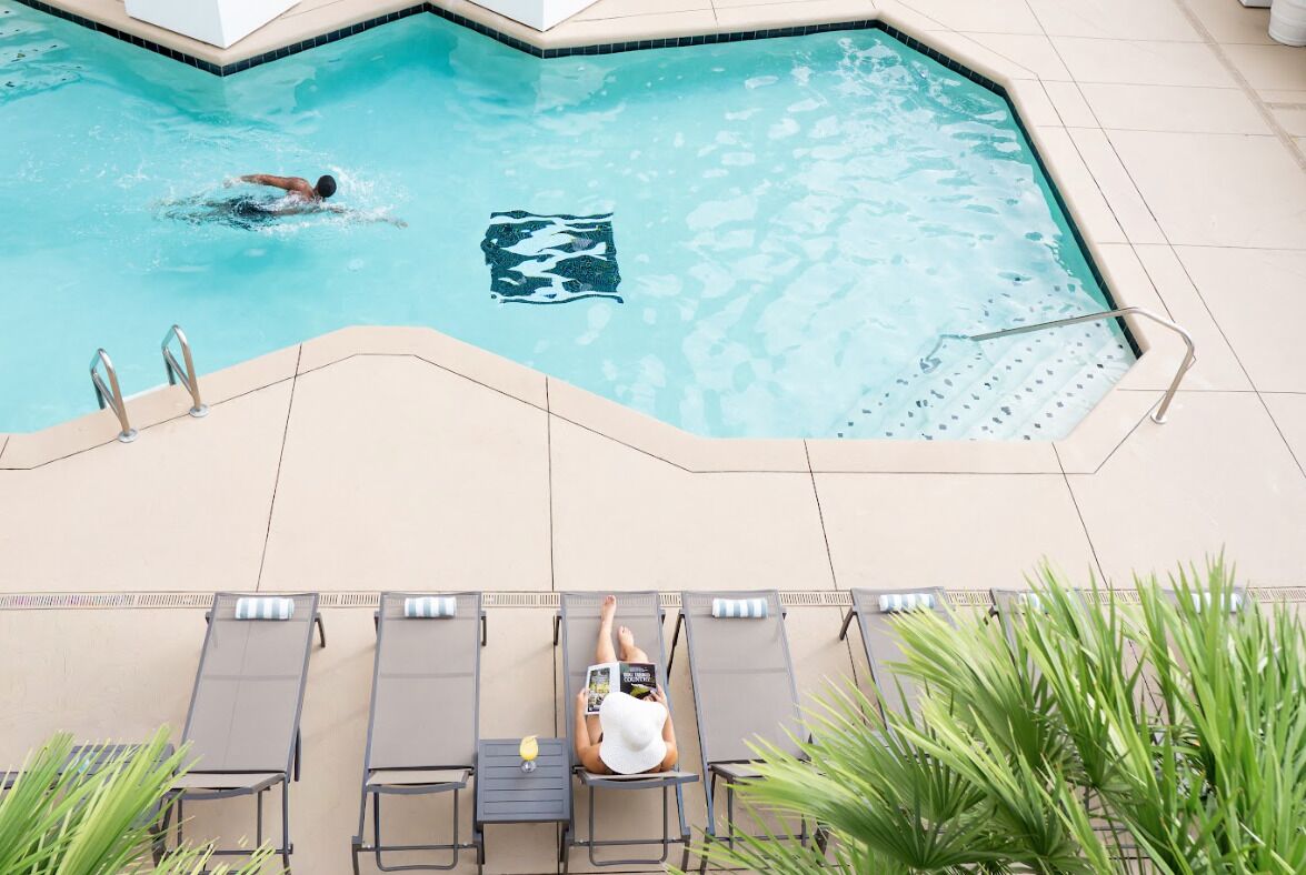 A swimming pool with a person swimming, lounge chairs, and a person reading a book.