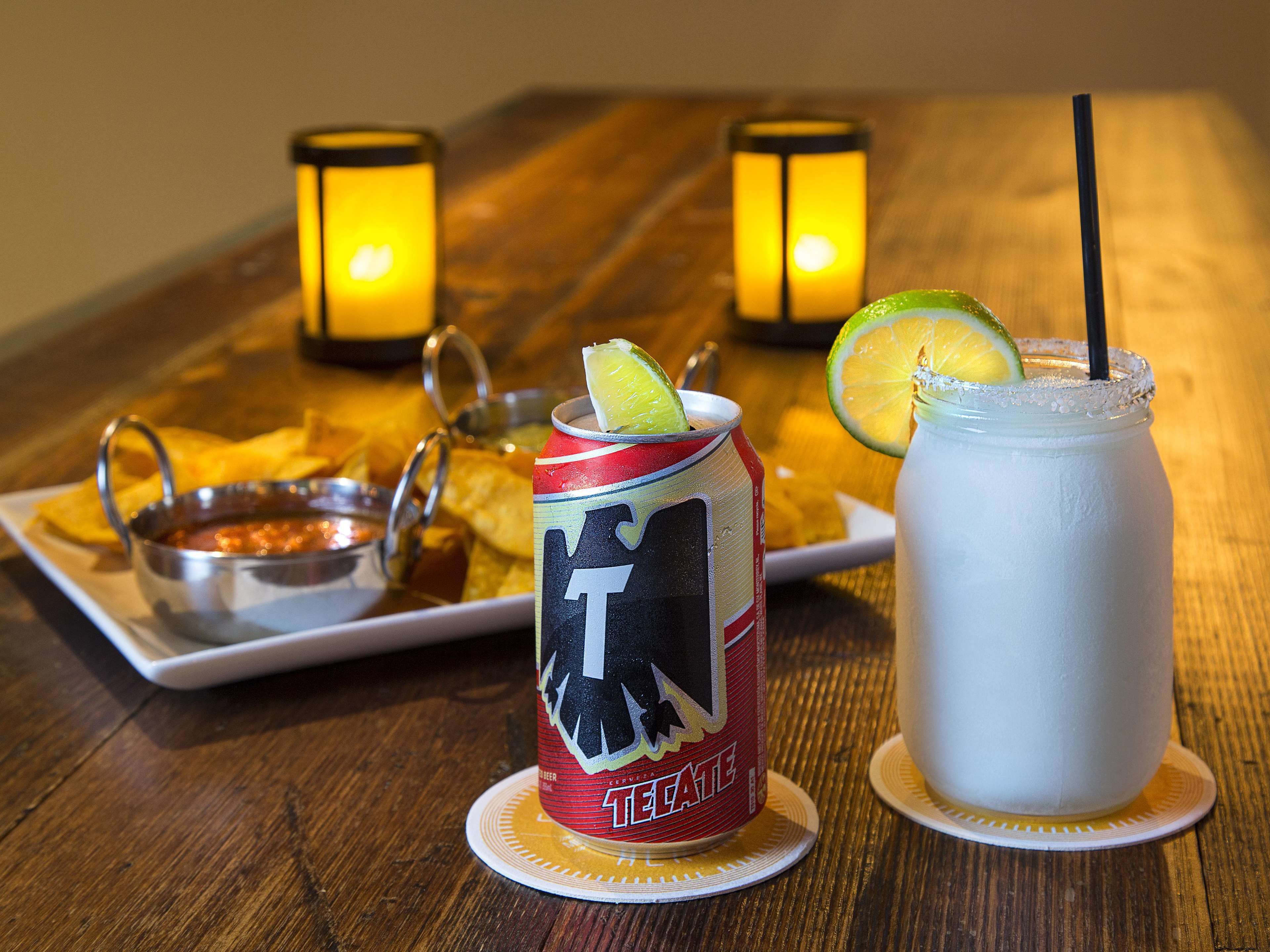 A can of Tecate beer, a lime wedge, and a white beverage with a straw on a wooden table.