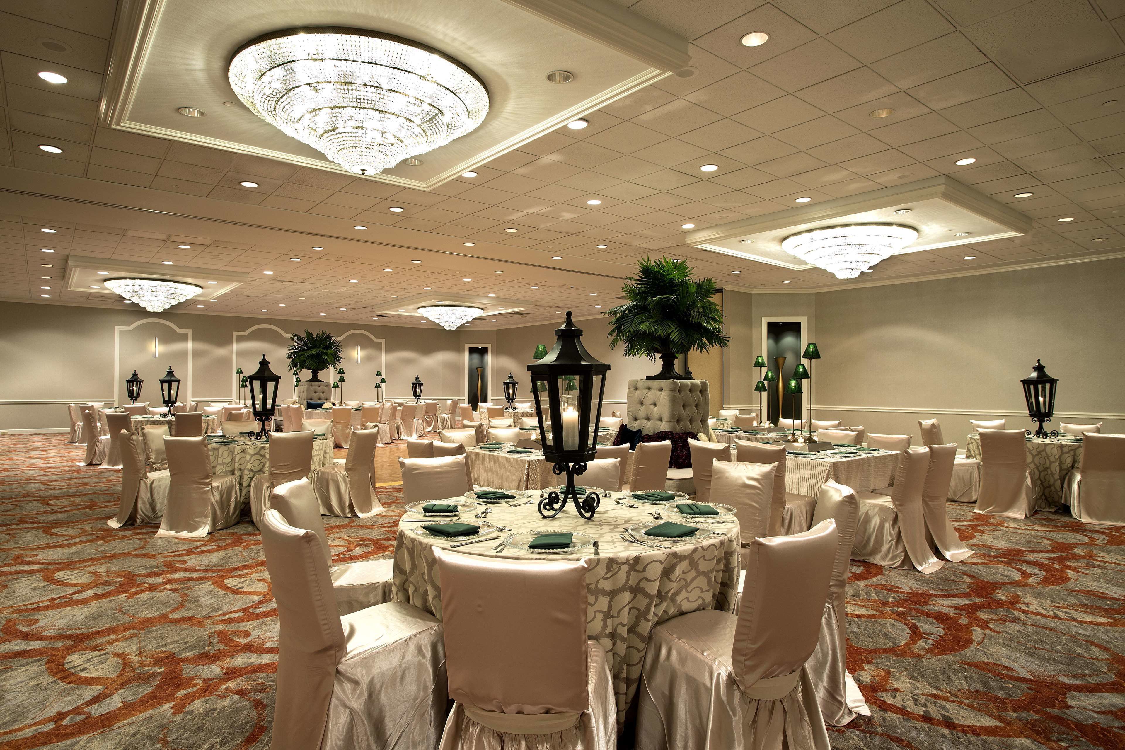 Une salle de banquet avec des tables rondes, des chaises blanches, des lustres et des plantes en pot.