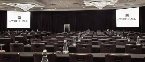 A conference room with rows of tables and chairs, large screens displaying 'The Whitehall' logo, and a chandelier on the ceiling.