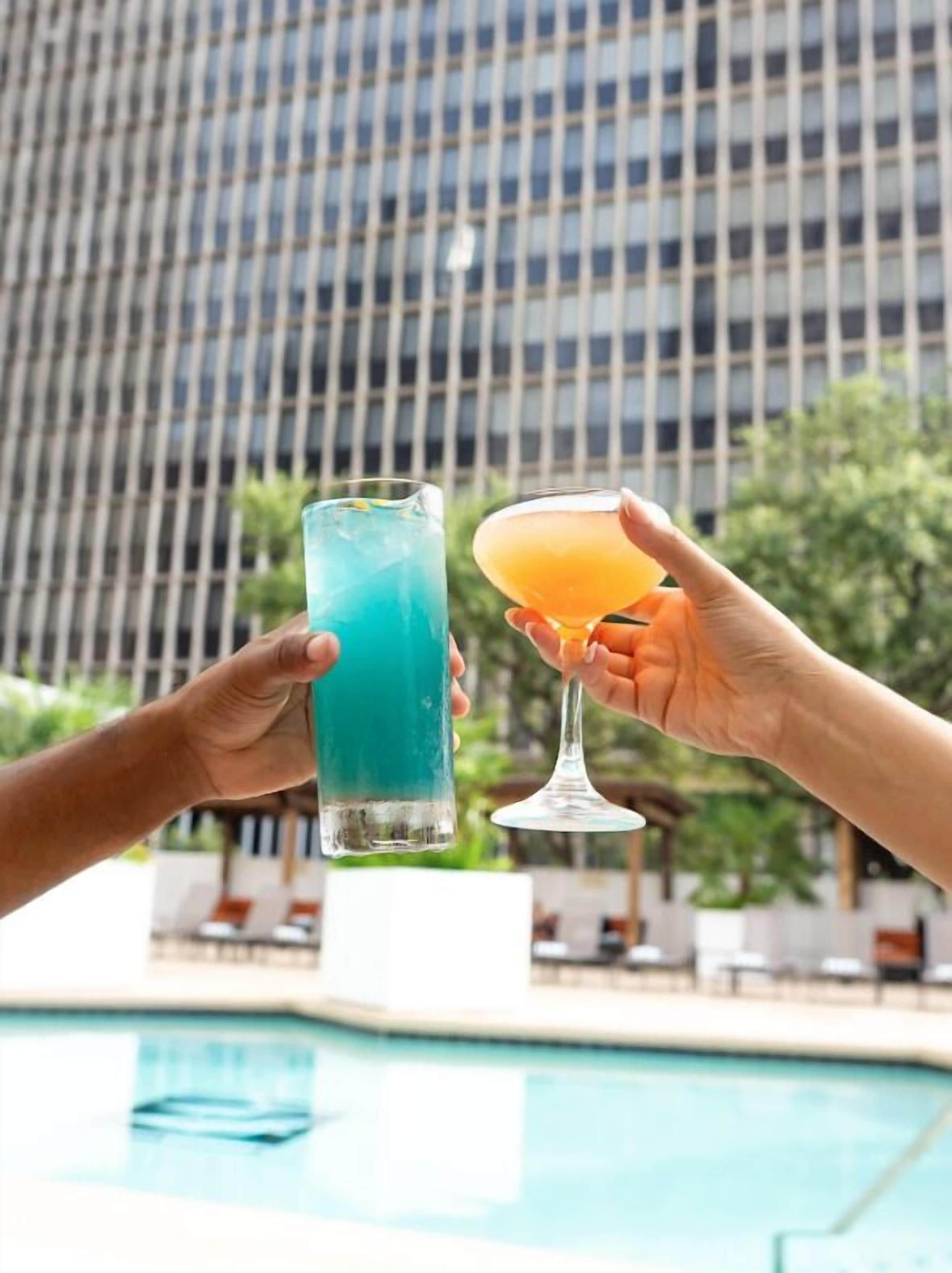Dos manos sosteniendo un vaso alto y una copa de cóctel con bebidas, frente a una piscina y un edificio con fachada enrejada.