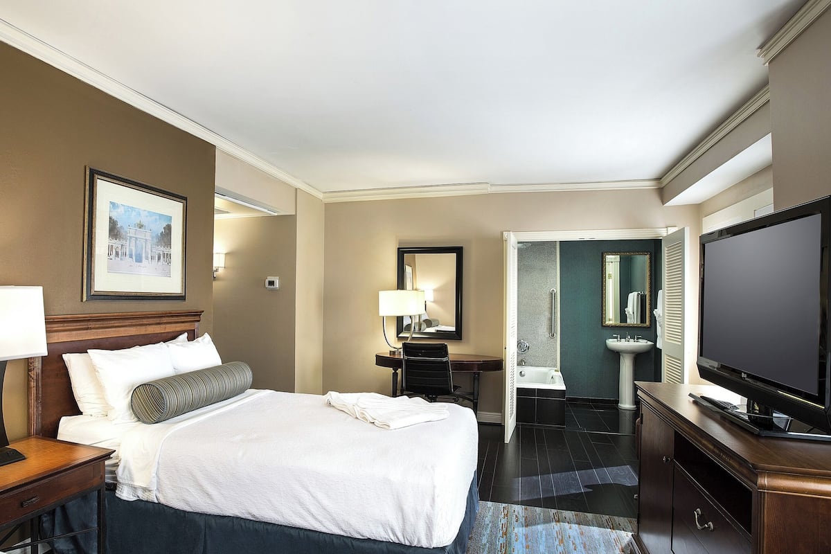 Ein Hotelzimmer mit einem großen Bett, einem Schreibtisch, einem Fernseher und einem Badezimmer mit Waschbecken und Spiegel.