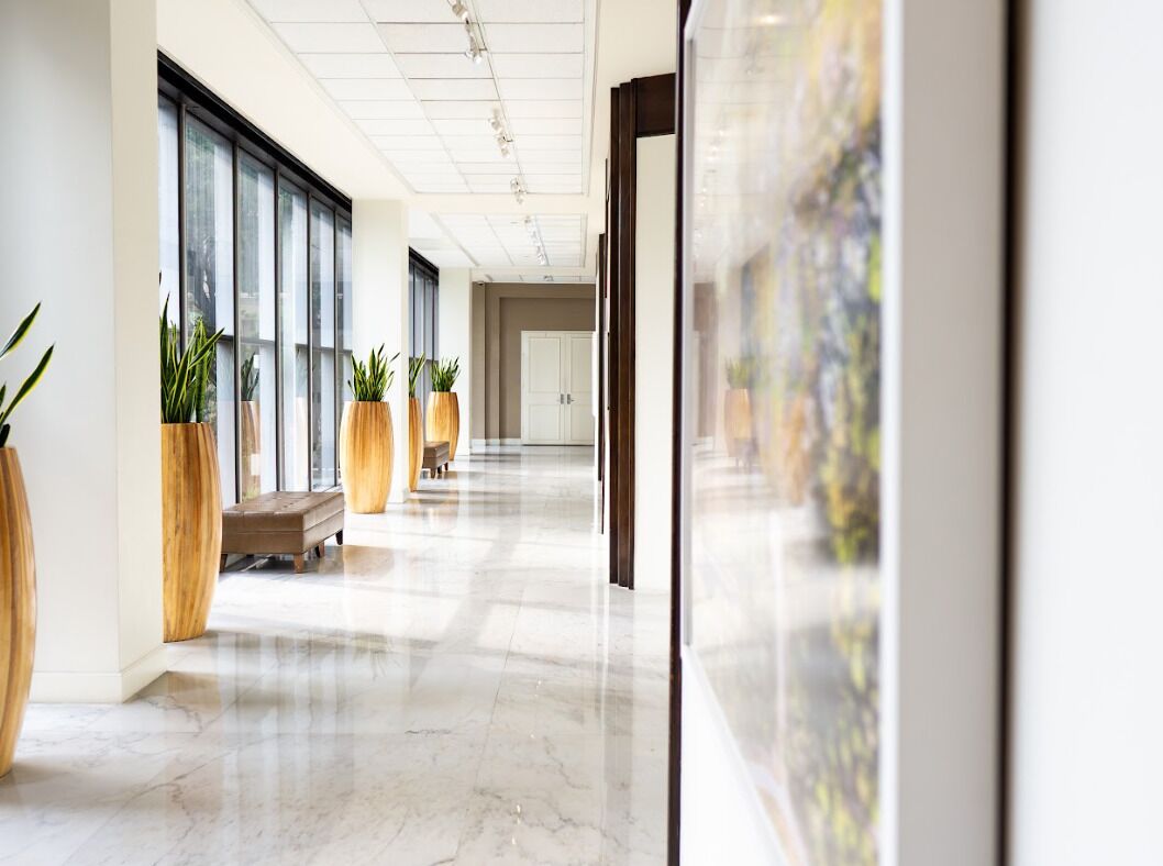 A modern hotel corridor with large windows, potted plants, and a marble floor.