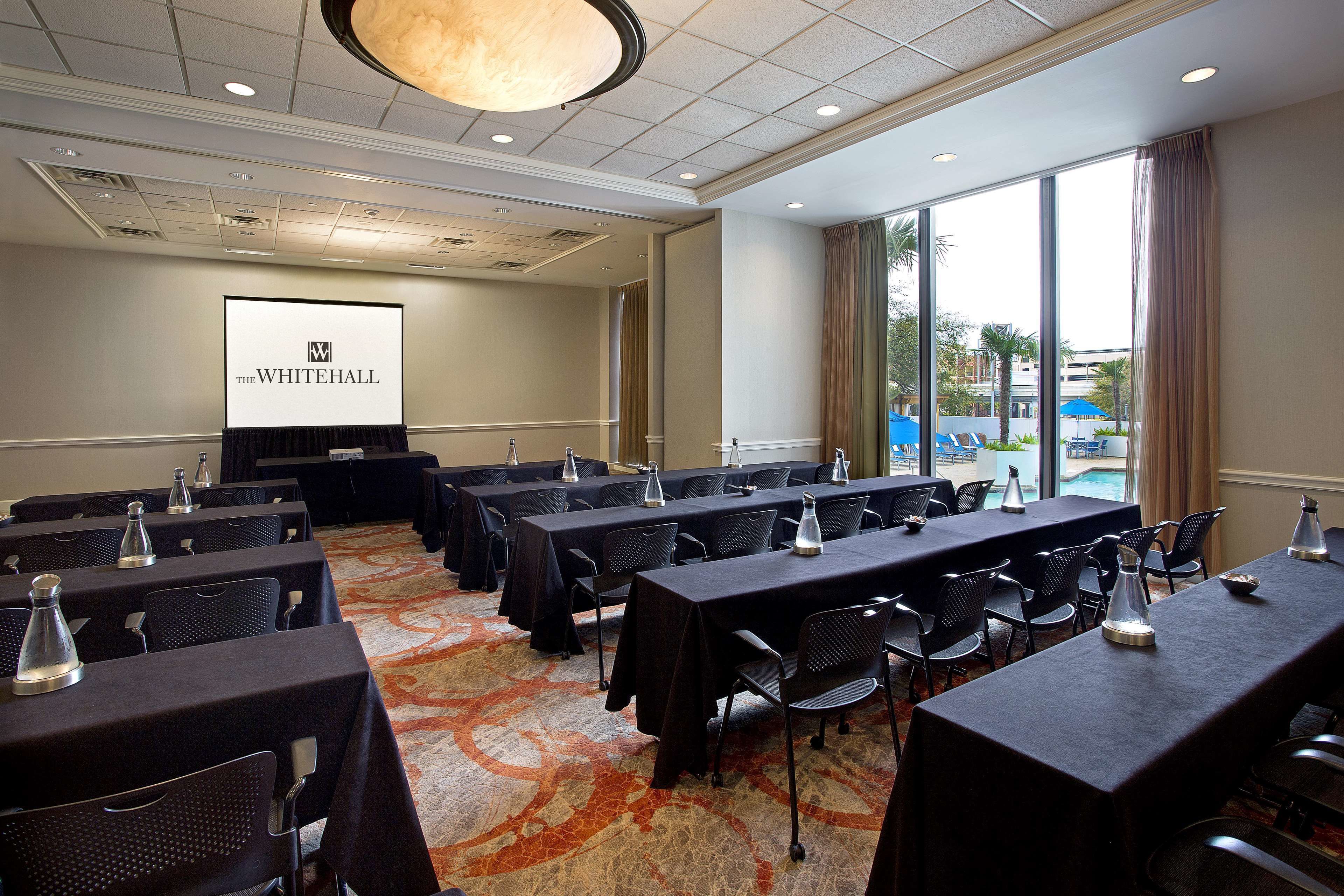 A conference room with multiple tables and chairs, a projector screen displaying "The Whitehall", and large windows offering a view of the outdoors.