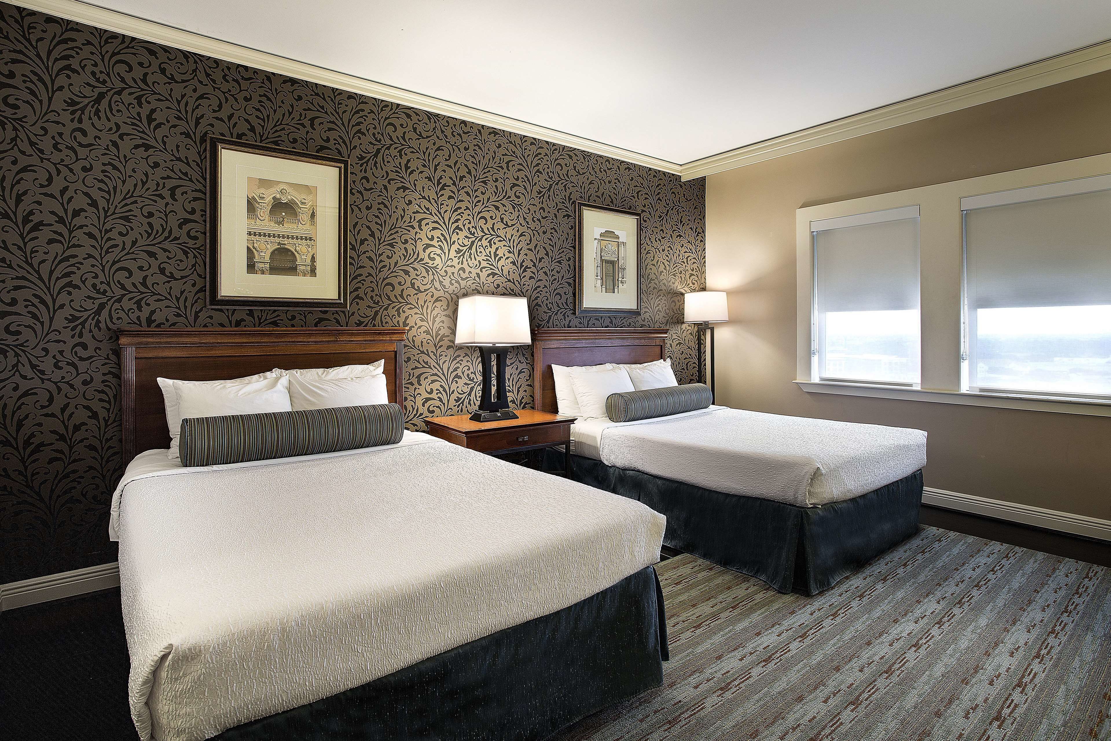 A hotel room with two beds, a nightstand with a lamp, framed pictures on the wall, and a window with a view.