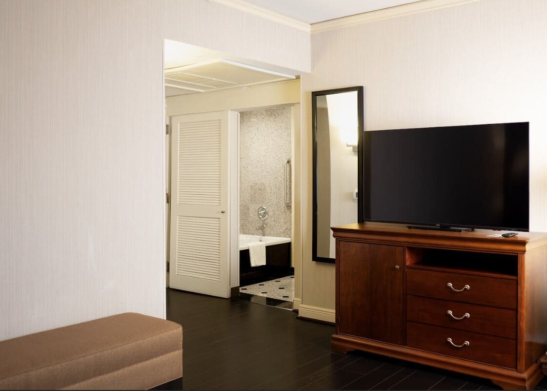 Habitación de hotel con un armario de madera, un televisor de pantalla plana, un banco, un baño con lavamanos y toalla, y una puerta que da a otra habitación.