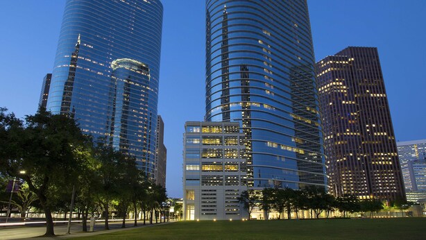 Paisagem urbana moderna com edifícios altos, incluindo um arranha-céu curvo com janelas iluminadas e um edifício retangular com fachada em grade.