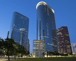 A modern cityscape with high-rise buildings, including a curved skyscraper with illuminated windows and a rectangular building with a grid-like facade. - The Whitehall Houston (Houston)