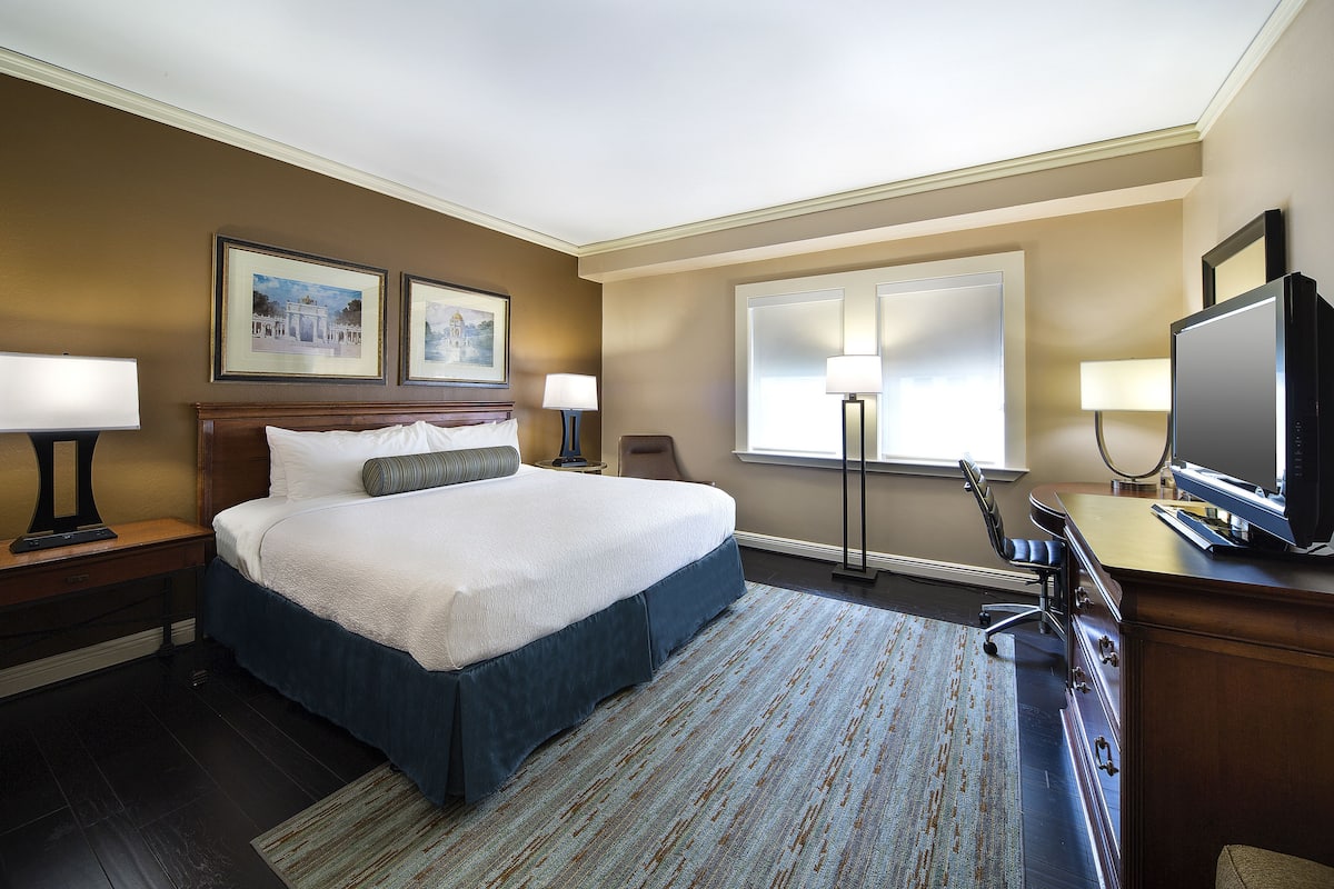 Ein Hotelzimmer mit einem großen Bett, zwei Nachttischlampen, einem Schreibtisch mit einem Computer und einem Fernseher.