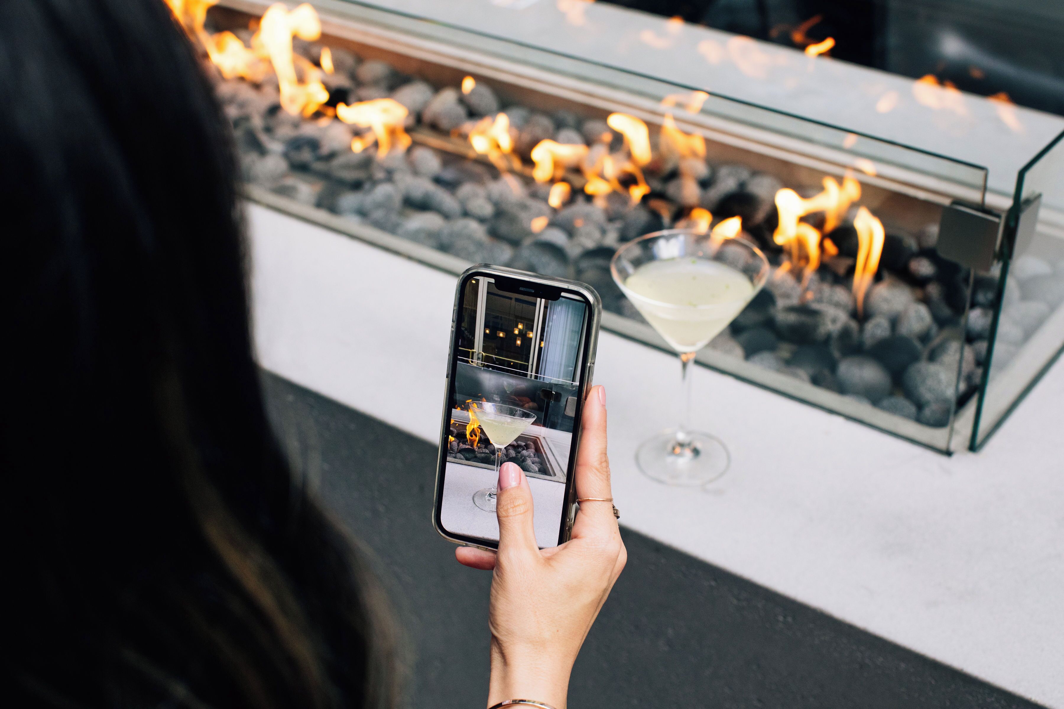 Una persona sosteniendo un teléfono inteligente, fotografiando un vaso de cóctel con un fuego encendido al fondo.