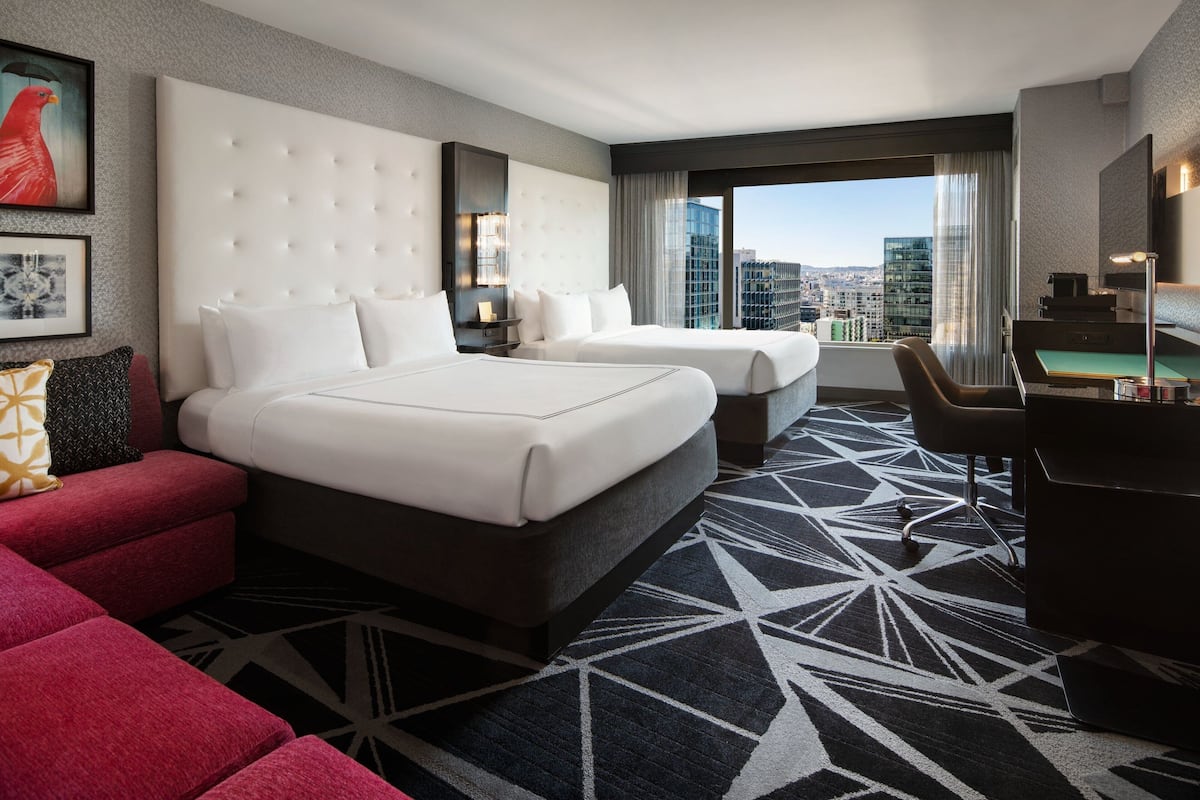 A hotel room with two beds, a desk, a red sofa, and a city view.