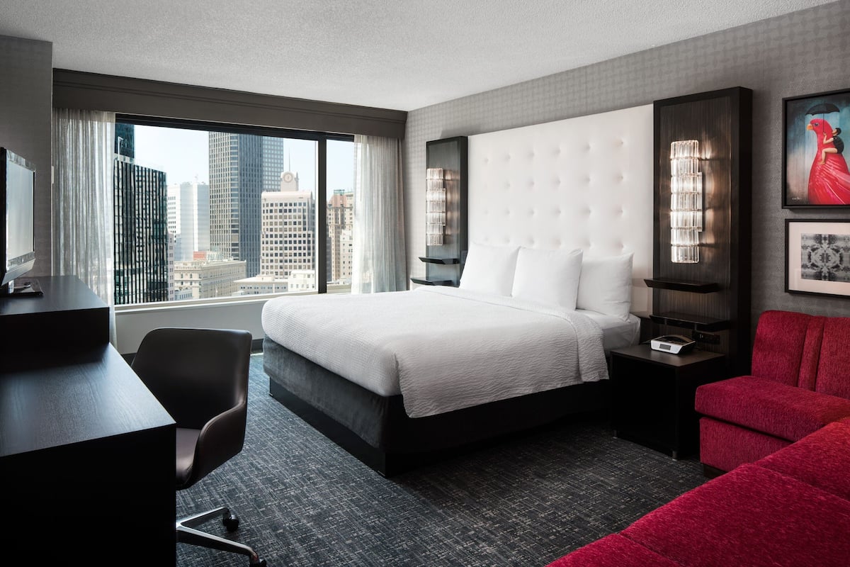 A hotel room with a large bed, a desk, a chair, a red sofa, a nightstand, and a city view.