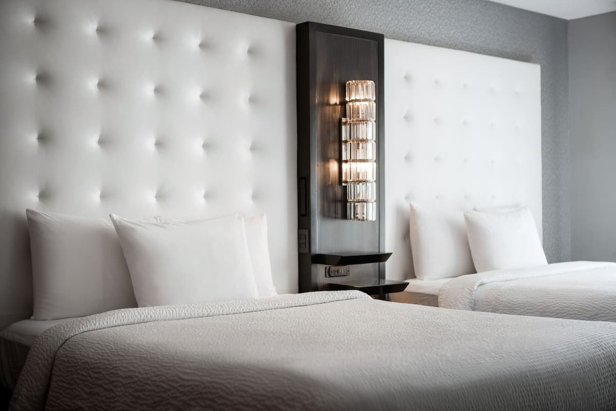 A hotel room with a tufted headboard, a bedside table with a lamp, and two beds with white bedding.
