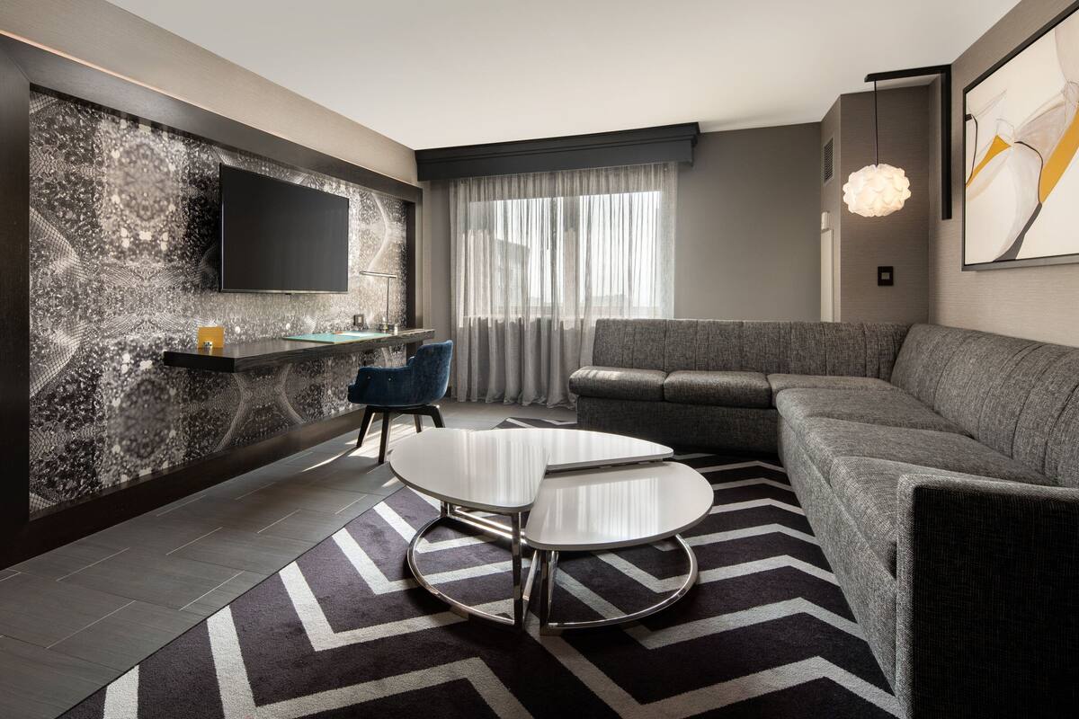 A modern living room with a sectional sofa, a flat-screen TV mounted on a textured wall, a small round table, and a patterned rug.