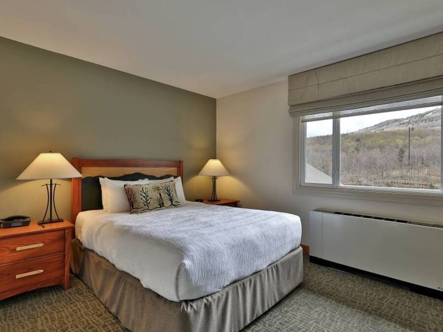 A hotel room with a bed, two bedside lamps, a nightstand, a window with a view of the outdoors, and a radiator.